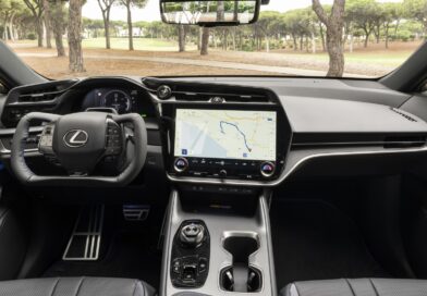 Inside a luxury car cockpit with a large touchscreen navigation system and digital gauge cluster behind the steering wheel.